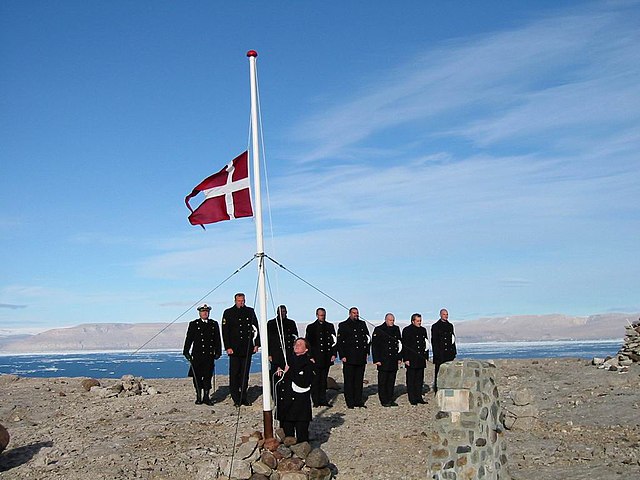 Eight Danish military officers watch as another hoists the Danish flag up a flagpole
