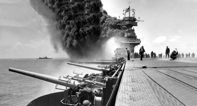 Scene on board the U.S. Navy aircraft carrier USS Yorktown (CV-5) during the Battle of Midway, shortly after she was hit by three Japanese bombs on 4 June 1942. Dense smoke is from fires in her uptakes, caused by a bomb that punctured them and knocked out her boilers. A man with hammer at right is probably covering a bomb entry hole in the forward elevator. Note arresting gear cables and forward palisade elements on the flight deck; CXAM radar antenna, large national ensign and YE homing beacon antenna atop the foremast; 12.7 cm/38, 12.7 mm caliber and 28 mm guns manned and ready at left. The heavy cruiser USS Astoria (CA-34) is visible on the left.
