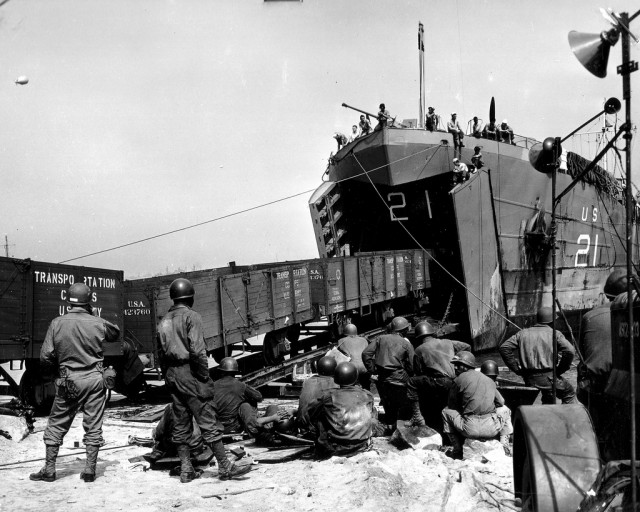 Le faible nombre de navires transport de trains va pousser à l'emploi de LST " spécialement aménagés en Ferry Train " (le faible tirant d'eau des LST leur permet d'accoster en se jouant des hauts-fonds) Le secteur nord du Terre-Plein des Mielles (à droite de la darse des Mielles) du port de Cherbourg est choisi, il pourra accueillir 6 LST Ferries Trains simultanément. Les trains pouvant grâce au nouveau réseau construit par les alliés, rejoindre le nud ferroviaire de la Gare Maritime. Travaux réalisés par le 342nd Eng General Service Rgt C'est d'ici que ces photos ont été prises, le 31 juillet 1944, le premier LST Ferry Train (LST-21) arrive et décharge le premier train, au terre-plein des Mielles sur la commune de Tourlaville. Pour réussir à décharger le train depuis les LST un système de bâti-rampe mobile est réalisé, la rampe mobile glisse jusqu'à arriver au niveau du pont inférieur du LST permettant l'ajustement final. Reportage : p012652, p012669, p012670, p012671, p012672, p012673, p012674, p012675, p012676, p012677 et p012681 Plusieurs wagons plateforme sortent du LST-21 Cette photo fait partie d'un reportage: http://www.flickr.com/photos/mlq/5529175303/