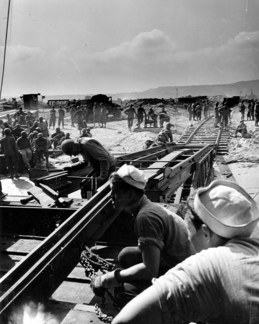 Le faible nombre de navires transport de trains va pousser à l'emploi de LST " spécialement aménagés en Ferry Train " (le faible tirant d'eau des LST leur permet d'accoster en se jouant des hauts-fonds) Le secteur nord du Terre-Plein des Mielles (à droite de la darse des Mielles) du port de Cherbourg est choisi, il pourra accueillir 6 LST Ferries Trains simultanément. Les trains pouvant grâce au nouveau réseau construit par les alliés, rejoindre le nœud ferroviaire de la Gare Maritime. Travaux réalisés par le 342nd Eng General Service Rgt C'est d'ici que ces photos ont été prises, le 31 juillet 1944, le  premier  LST Ferry Train (LST-21) arrive et décharge le premier train, au terre-plein des Mielles sur la commune de Tourlaville. Pour réussir à décharger le train depuis les LST un système de bâti-rampe mobile est réalisé,  la rampe mobile glisse jusqu'à arriver au niveau du pont inférieur du LST permettant l'ajustement final. Voir una autre vue page 9 de ce document: http://www.scribd.com/doc/279812/Railroads-WWII-ETO Reportage : p012652, p012669, p012670, p012671, p012672, p012673, p012674, p012675, p012676, p012677 et p012681 Installation de la rampe de raccordement. Cette photo fait partie d'un reportage: http://www.flickr.com/photos/mlq/5529175303/