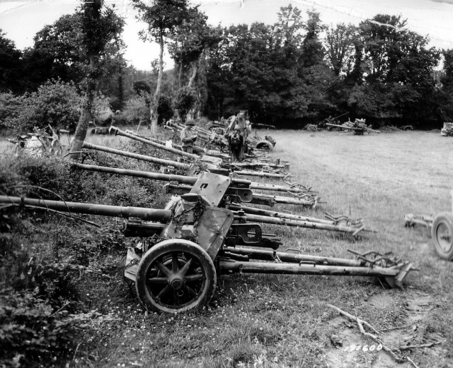 Dans un champ, du matériel allemand stocké, pièces d'artillerie. Dans ce site, Cote: 13 Num 510 en date du 13 juillet 1944 http://recherche.archives.manche.fr/?id=recherche_documents_figures le zoom est plus fort et le tirage plus large ce qui permet une meilleure analyse. Voir ici le repérage des pièces : http://www.flickr.com/photos/mlq/6219734677/in/photostream Alignés le long d'une haie : six 7,5 cm Pak 40 , deux 10,5 cm leFH 18/40, et différents autres canons de Flak ou antichars. Un GI prend des notes sur un carnet, derrière lui un projecteur 150cm Flakscheinwerfer 34. Voir ici en milieu de page: http://www.dzwi.de/60cmScheinwerfer.htm Photo prise dans un dépôt US près d'Isigny-sur-Mer ; localisation, après le croissement de la D196 et la D203 en direction de Lison, à l'est du hameau de la Madeleine. voir ici: http://www.flickr.com/photos/mlq/5776184661/in/photostream L'auteur de ce livre: http://www.priceminister.com/offer/buy/6201586/John-S-Allsup-L-enfer-Du-Bocage-Hedgerow-Hell-Livre.html John S. Allsup, 1st platoon, Co A,175th Inf., 29th US ID (il a débarqué à Omaha, le 7 juin 1944). Date cette photo du 13 juillet 1944. Reportage de 6 photos sur ce parc de matériel allemand : 4 repérées : p012031, p012032, p013086 et p013441. Et deux, sous réserve : p012028 et p.012034. http://www.flickr.com/photos/mlq/5786951602/in/photostream Selon ce site : une autre vue de ce dépôt de matériel allemand http://recherche.archives.manche.fr/?id=recherche_documents_figures Cote: 13 Num 4555 Date : 25 juillet 1944