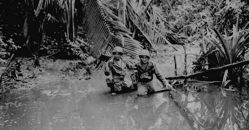 Sgt. Carl Weinke and Pfc. Ernest Marjoram, Signal Corps cameramen, wading through stream while following infantry troops in forward area during invasion at a beach in New Guinea.