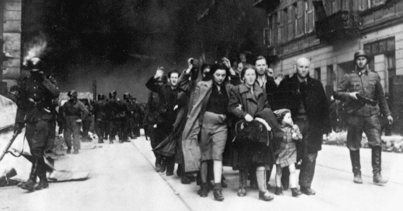 Ghetto buildings burning while German soldiers march Jewish people away. 