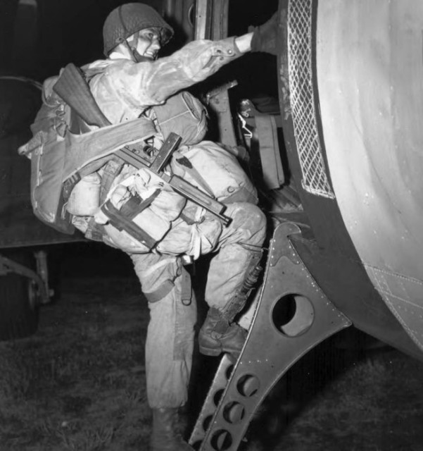 Fully Equipped paratrooper armed with a Thompson submachine gun M1, climbing into a transport plane to go to France as the invasion of Normandy gets under way. At approximately 0200, 6 June 1944, men of two U. S. airborne divisions, as well as elements of a British airborne division, were dropped in vital areas to the rear of German coastal defenses guarding the Normandy beaches from Cherbourg to Caen. By dawn 1,136 heavy bombers of the RAF Bomber Command had dropped 5,853 tons of bombs on selected coastal batteries lining the Bay of the Seine between Cherbourg and Le Havre. Image and caption credit: Center of Military History. U.S. Army (Image)
