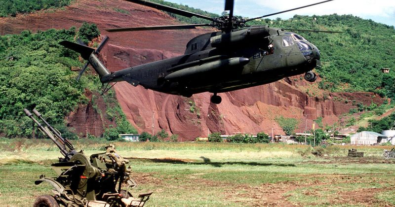 A CH-53 Sea Stallion helicopter hovers above the ground near a Soviet ZU-23 anti-aircraft weapon prior to picking it up during Operation URGENT FURY.