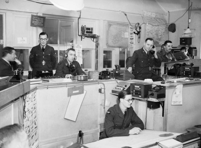 Interior of the Sector 'G' Operations Room at Duxford, Cambridgeshire. The callsigns of fighter squadrons controlled by this Sector can be seen on the wall behind the operator sitting third from left. The Controller is sitting fifth from the left, and on the extreme right, behind the Army Liaison Officer, are the R/T operators in direct touch with the aircraft. [© IWM (CH 1401)]