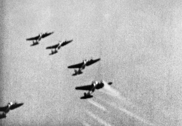 A still from camera-gun film taken from a Supermarine Spitfire Mark I of No. 609 Squadron RAF, flown by by Pilot Officer J D Bisdee, as he dives on a formation of Heinkel He 111s of KG 55 which have just bombed the Supermarine aircraft works at Woolston, Southampton. The rearmost aircraft of the leading 'staffel' receives a burst of machine gun fire from Bisdee, as shown by the streaks of light from the tracer bullets. Its port engine is also on fire. [© IWM (CH 1827)]