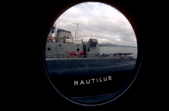 A view through a porthole aboard the large harbor tub PUSHMATAHA (YTB 830) of the nuclear-powered attack submarine ex-USS NAUTILUS (SSN 571) and large harbor tug SKENANDOA (YTB 835).