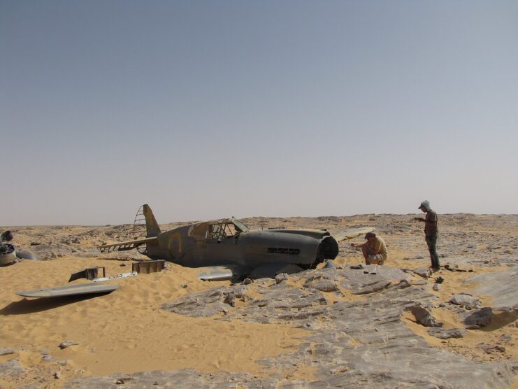 Two men standing around a Curtiss P-40 Kittyhawk half-buried in the sand