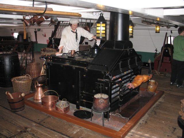 A sailor standing over a Royal Navy stove, onboard HMS Trincomalee. These stoves were designed to mass produce meals, being able to hold multiple cooking pots, and even a rotisserie. Image Source: Wikimedia Commons/ By Ian Petticrew, CC BY-SA 2.0, https://commons.wikimedia.org/w/index.php?curid=8732582