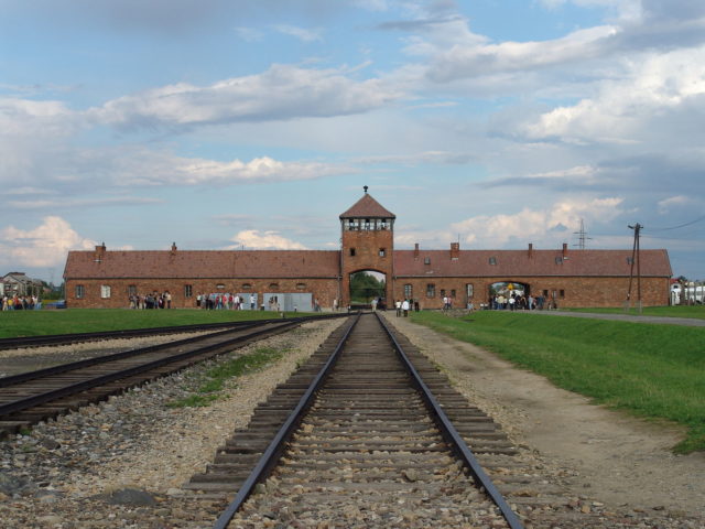 The main gate at the former German Nazi concentration camp of Auschwitz II (Birkenau). Note that this is inside the camp looking back from the loading ramp to the “Gate of Death”. By Michel Zacharz AKA Grippenn – CC BY-SA 2.5