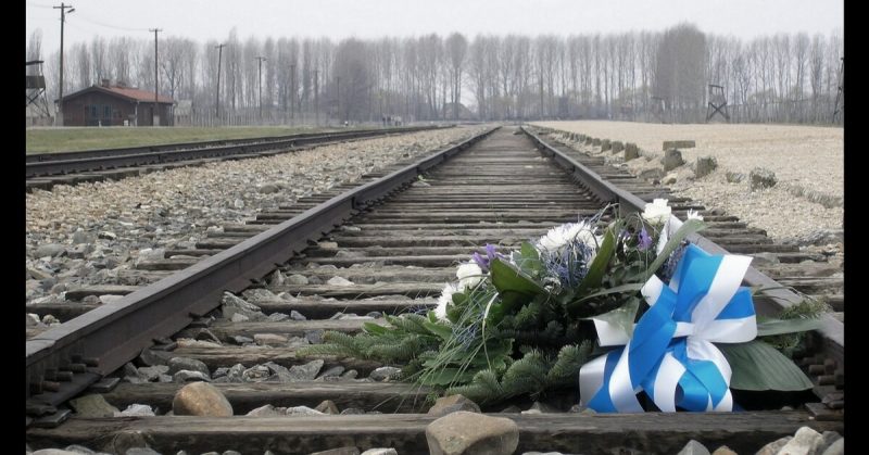 A wreath laid on the rail tracks at Auschwitz, 2007. <a href=https://commons.wikimedia.org/wiki/File:KZ_Auschwitz-Birkenau,_Bahngleise_der_Entladerampe,_Blumen_zum_Gedenken.jpg>Photo Credit</a>