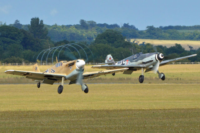 Two 109s take-off at Duxford. Both are Post-war Spanish built Hispano Buchons and both also flew from Duxford in the late 1960’s during production of the ‘Battle of Britain’ film. To the right is ‘D-FWME’ which is now fitted with a DB605 engine and is effectively a BF109G-4. Left is ‘G-AWHE’ in a desert scheme. This Buchon is still Merlin powered and is generally operated by ARCo. They are seen getting airborne for the ‘Balbo’ at the 2015 Flying Legends Airshow. Duxford, Cambridgeshire, UK. 12-7-2015. Photo: Alan Wilson from Stilton, Peterborough, Cambs, UK / CC BY-SA 2.0