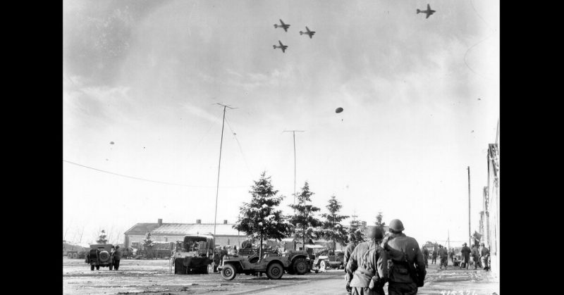 Photo taken by a U.S. Army Signal Corps photographer on 26 December 1944 in Bastogne, Belgium as troops of the 101st Airborne Division watch C-47s drop supplies to them. Jeeps and trucks are parked in a large field in the near distance.