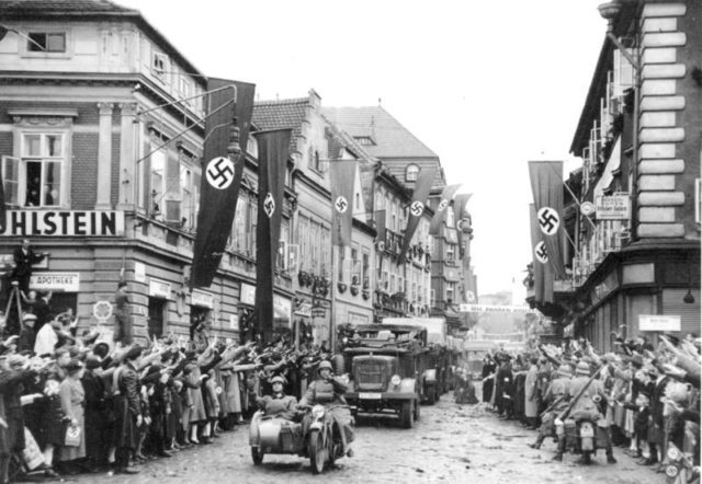 German mechanized troops enter Saaz, Sudetenland. The streets are decorated with swastika flags and banners. 9.10.1938. Bundesarchiv – CC BY-SA 3.0 de