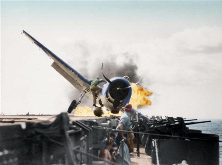 Crash landing of F6F-3, Number 30 of Fighting Squadron Two (VF-2), USS Enterprise, into the carrier’s port side 20mm gun gallery, 10 November 1943. Lieutenant Walter L. Chewning, Jr., USNR, the Catapult Officer, is climbing up the plane’s side to assist the pilot from the burning aircraft. The pilot, Ensign Byron M. Johnson, escaped without significant injury. Enterprise was then en route to support the Gilberts Operation. Note the plane’s ruptured belly fuel tank. Paul Reynolds / mediadrumworld.com