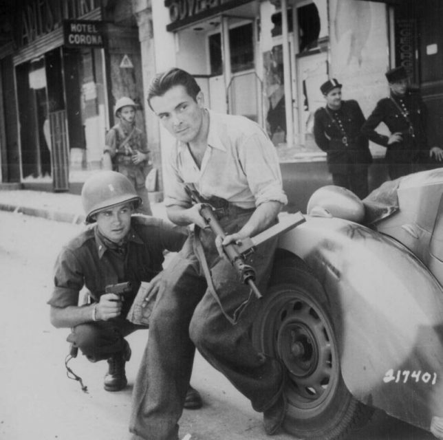 An American officer with a colt 1911 and a French partisan with a Sten sub-machinegun crouched behind a car during a street fight in a French city, June 1944.