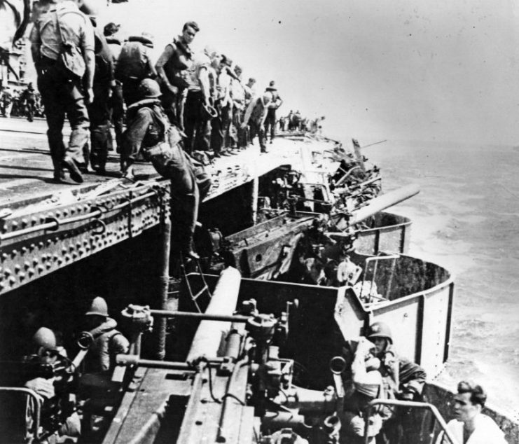 Damage in the port forward 5-inch gun gallery on USS Lexington (CV-2), from a Japanese bomb that struck near the gallery’s after end during the Battle of the Coral Sea, 8 May 1942. View looks aft, with the ship’s number two 5/25 gun in the foreground, still manned and in operation. Number four 5/25 gun is immediately beyond, trained out to port and aft.