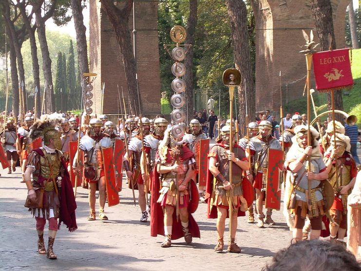Modern reenactors parade with replicas of various legionary standards. From left to right: signum (spear-head type), with four discs; signum (wreathed-palm type), with six discs; imago of ruling emperor; legionary aquila; vexillum of commander (legatus) of Legio XXX Ulpia Victrix, with embroidered name and emblem (Capricorn) of legion. Photo: Marten253 / CC BY-SA 3.0