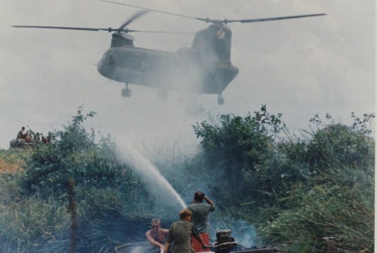 Three US Army servicemen spraying herbicide along a river bank while a helicopter hovers overhead