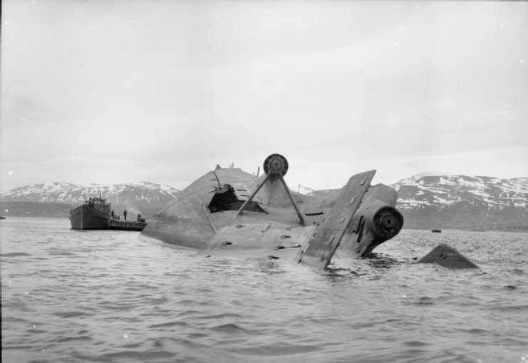 The German battleship Tirpitz, lying capsized in Tromso fjord, attended by a salvage vessel. The already damaged ship was finally sunk in a combined daylight attack by Nos. 9 and 617 Squadrons RAF on 12 November 1944, (Operation Catechism). The hole in the hull by the starboard propeller shaft was cut by the Germans to allow access to salvage crews.