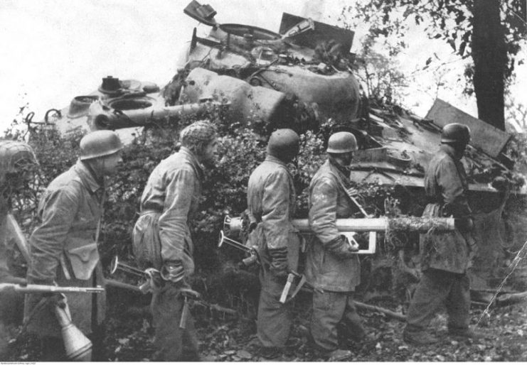 German Fallschrimjagers passing by a destroyed Sherman tank, Normandy, 1944. Note Panzerschreck and Panzerfaust anti-tank weapons.