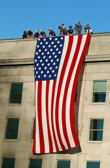 Military service members render honors as fire and rescue workers unfurl a huge American flag over the side of the Pentagon during rescue and recovery efforts following the 11 Sept. terrorist attack. U.S. Navy photo by Michael W. Pendergrass.