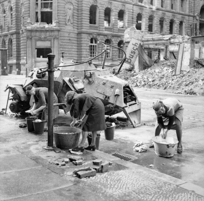 German women doing their washing at a cold water hydrant in a Berlin street, a knocked out German scout car stands beside them.