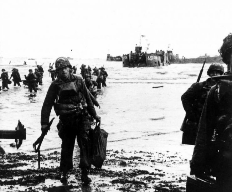 Carrying full equipment, American assault troops move onto Utah Beach. Landing craft, in the background, jams the harbor.