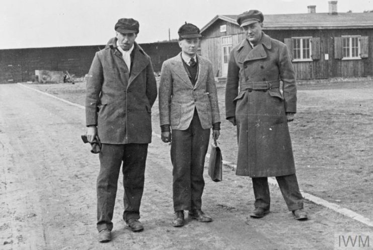 A full-length portrait of three Prisoners of War disguised as German civilians at Stalag Luft III, Sagan. Behind them can be seen one of the camp huts. IWM