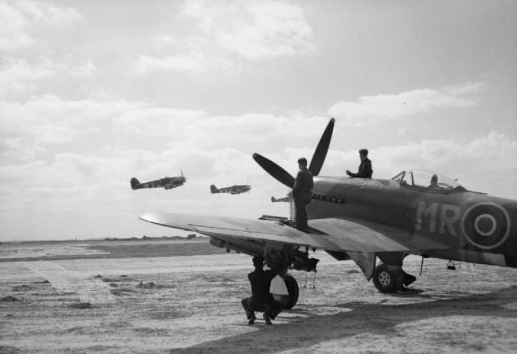 A section of Hawker Typhoon Mark IBs of No. 175 Squadron RAF take off from B5/Le Fresne Camilly, Normandy, watched by armorers at work on another Typhoon.