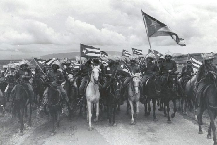 1959 Cuban Revolution entitled “La Caballería” (The Cavalry). The image shows a group of Fidel Castro’s July 26 Movement rebels mounted on horses and brandishing Cuban flags.