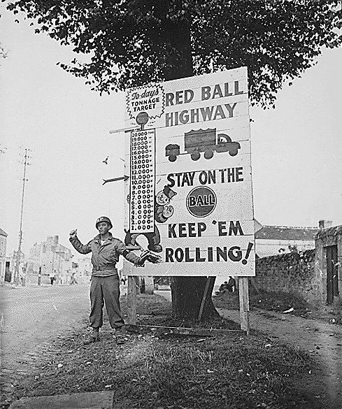 Corporal Charles H. Johnson of the 783rd Military Police Battalion, waves on a “Red Ball Express” motor convoy rushing priority materiel to the forward areas, near Alenon, France.