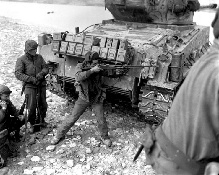 Taking cover behind their tank escort, one man of this ranger patrol of the 5th RCT, U.S. 24th Infantry Division, uses his BAR to return the heavy Chinese Communist small arms and mortar fire. On the left, another soldier uses a field radio to report the situation to headquarters.