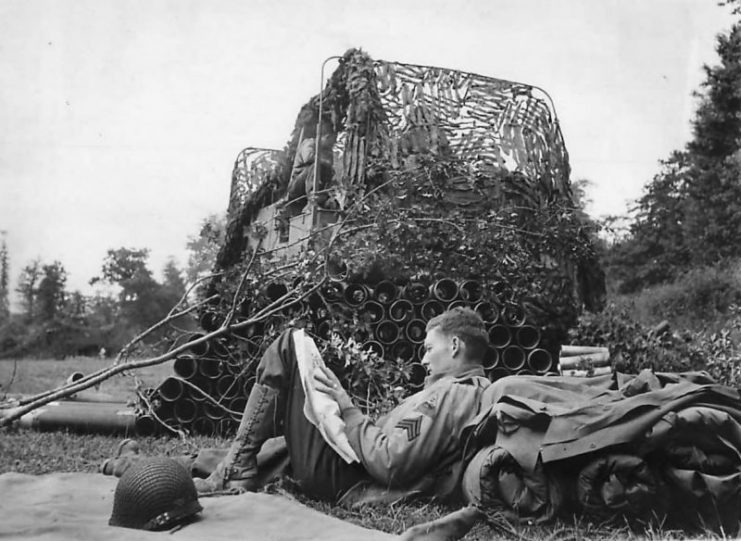 Soldier reads newspaper by camouflaged M7 near Carentan, 1944