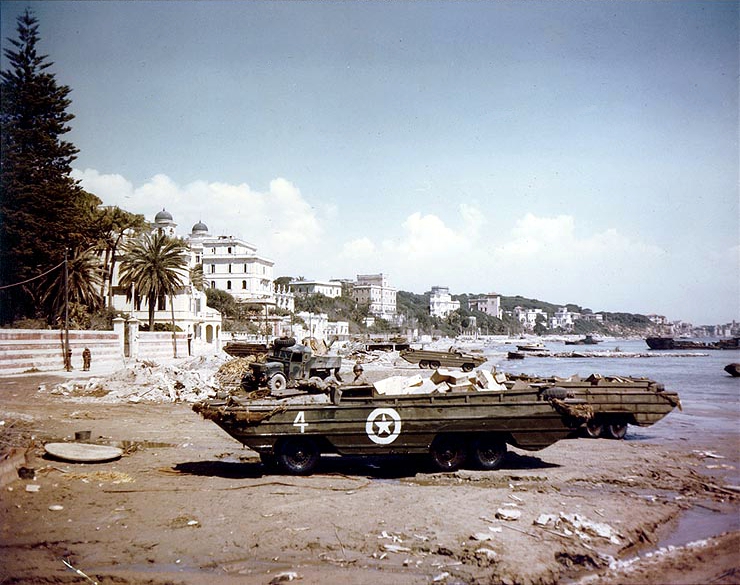 U.S. Army DUKW amphibious trucks transporting cargo across the beach at Anzio, 15 April 1944. USS LCT-33 is partially visible in the right background.