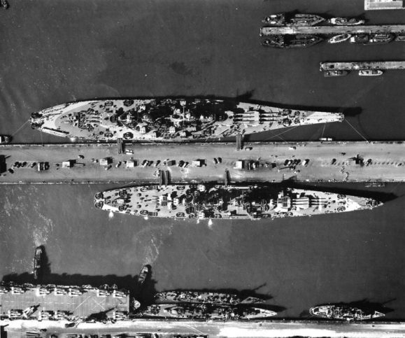 Aerial view of warships at the base piers of Norfolk Naval Base, Virginia (USA), circa August 1944. Among them are: the battleship USS Missouri (BB-63), the largest ship; the battlecruiser USS Alaska (CB-1), on the other side of the pier; the escort carrier USS Croatan (CVE-25), and two destroyers, a Fletcher-class destroyer at the pier and a Clemson/Wilkes-class-destroyer moored outboard.