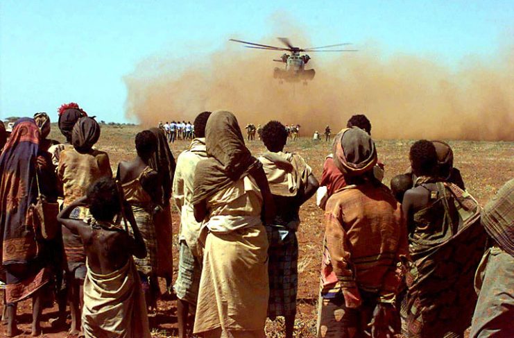 The camera shoots past some Somalis from the village of Maleel as they watch a US Marine CH-53 Sea Stallion deliver a sling load of wheat donated by the people of Australia.