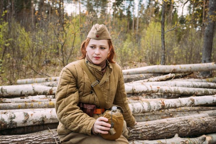Pribor, Belarus – April 23, 2016: Young Woman Re-enactor Dressed As Russian Soviet Infantry Soldier Of World War II Sitting On Wooden Logs, And Drinks Water From A Flask In Forest