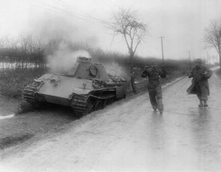 A German prisoner is marched past a burning Nazi tank on a Belgian road. Sgt. Bernard Cook of Los Angeles, Calif of the 165th Photo Company, 1st US Army, walks behind the captured soldier.