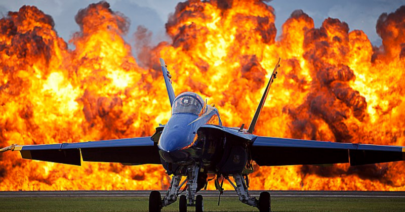 A wall of fire erupts behind a U.S. Navy F/A-18 Hornet aircraft