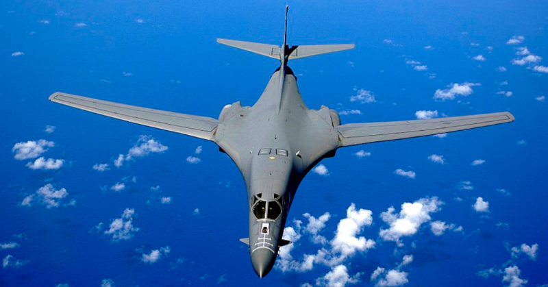 Top view of B-1B in-flight with white clouds scattered underneath. Its wings are swept fully forward as the gray aircraft flies over the ocean.