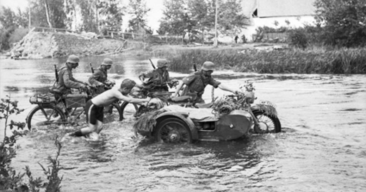 Troops pushing a motorcycle through a river. By Bundesarchiv, Bild CC-BY-SA