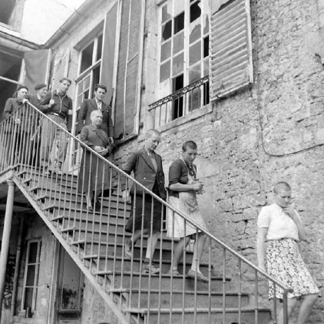 Four mowed women go down an outside staircase, preceding four civilians, one of whom is armed with a rifle. Photo: PhotosNormandie / CC BY-SA 2.0