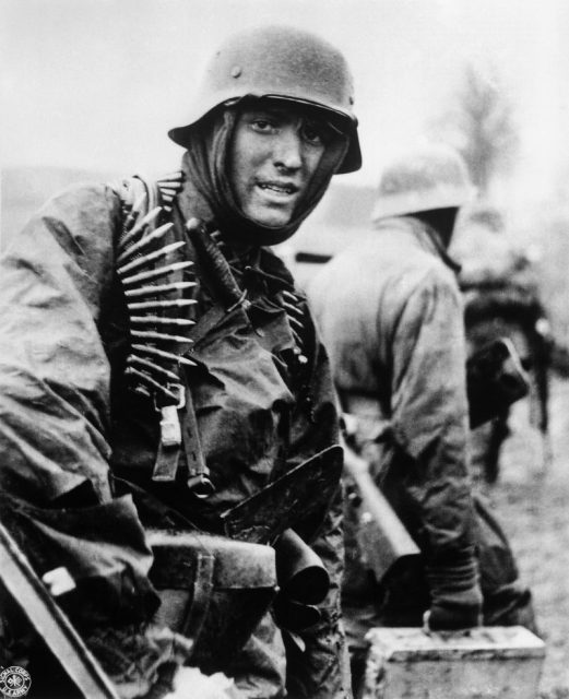 A German soldier, heavily armed, carries ammunition boxes forward with companions in territory taken by their counter-offensive in Ardennes.