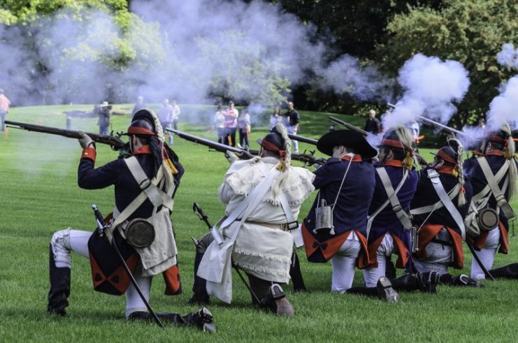 Colonial sharpshooters fire muskets on a mock battlefield, while spectators look on, at a reenactment of the American Revolutionary War (1775-1783).