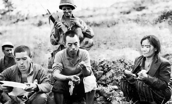 Two surrendered Japanese soldiers with a Japanese civilian and two US soldiers on Okinawa. The Japanese soldier on the left is reading a propaganda leaflet.