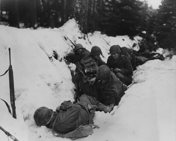US Soldiers crouch in a snow-filled ditch, taking shelter from a German artillery barrage in the Krinkelter woods.