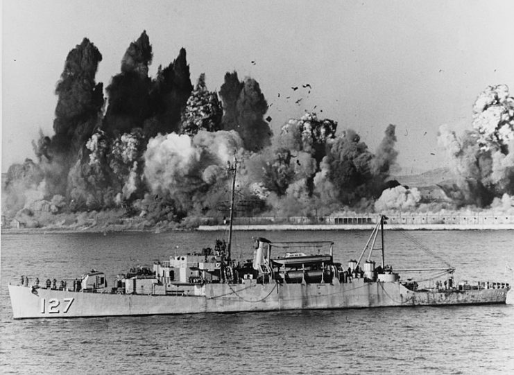 During the evacuation of Hŭngnam, Korea, on 24 December 1950, the U.S. Navy high-speed transport USS Begor (APD-127) stands offshore, ready to embark the last UN landing craft, as demolition charges wreck Hŭngnam’s port facilities.