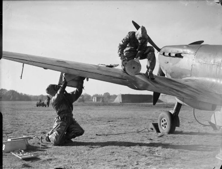 Armourers of No. 403 Squadron RCAF, wearing anti-gas equipment, re-arm the cannons and machine guns of Supermarine Spitfire Mark V
